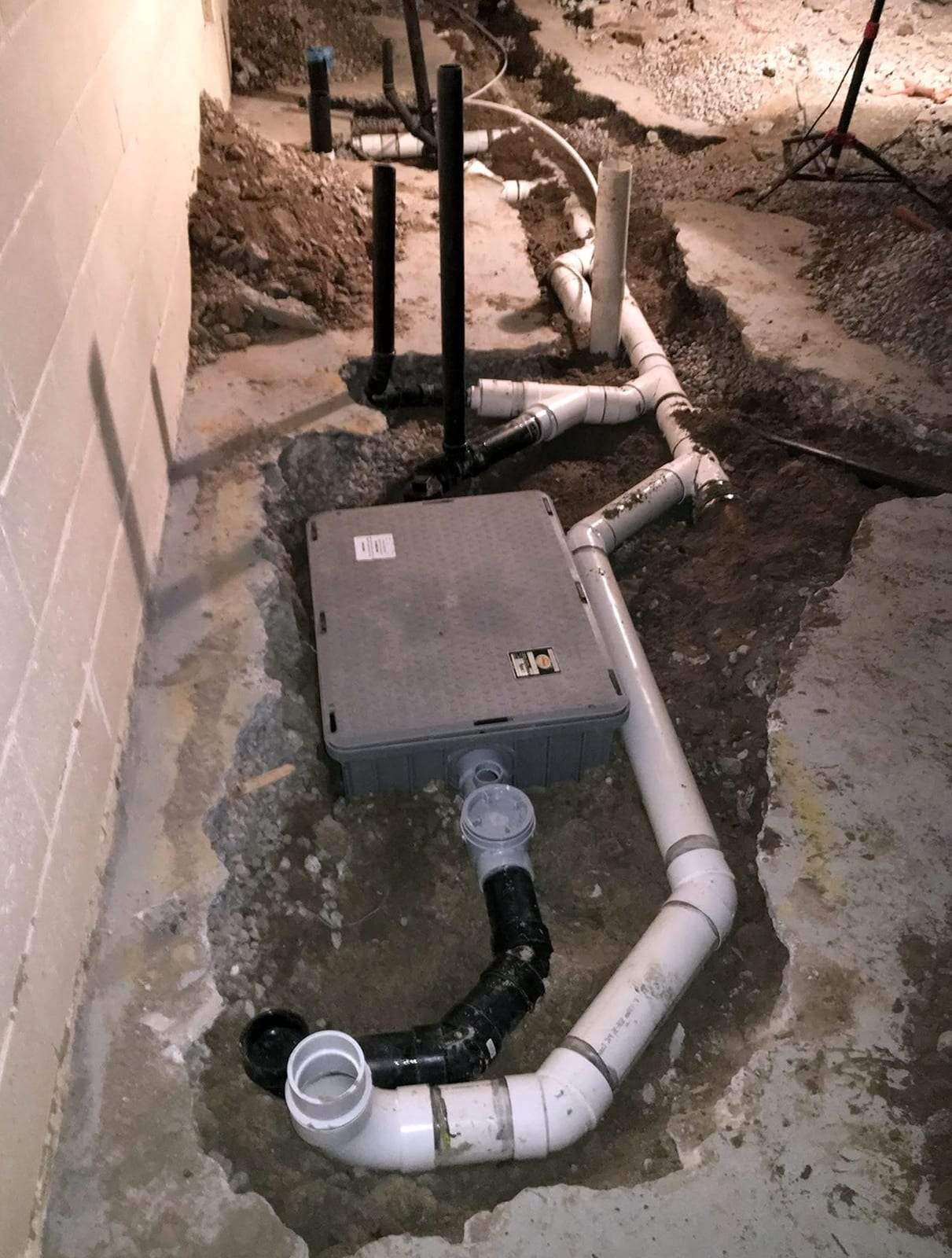 New PVC drain lines and an ejector basin with a gray lid set in an opened trench in a basement floor