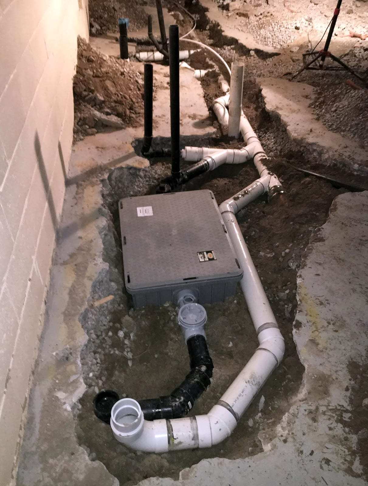 New PVC drain lines and an ejector basin with a gray lid set in an opened trench in a basement floor
